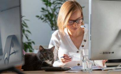 Eine Frau sitzt vor dem PC-Bildschirm und liest handschriftliche Notizen, neben ihr sitzt eine Katze auf dem Schreibtisch