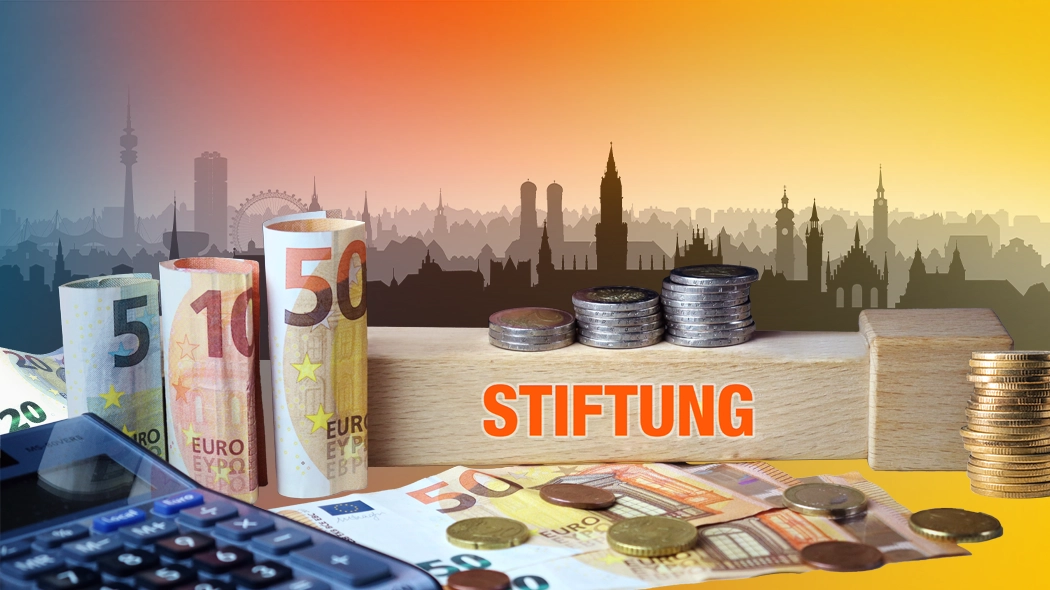 Symbolbild zum Thema Stiftung: Euro-Banknoten und Münzen liegen neben einem Taschenrechner auf einem Tisch. Auf einem Holzblock mit dem Schriftzug ‚STIFTUNG‘ sind Münzen gestapelt. Im Hintergrund ist die Silhouette einer Stadt mit Sonnenuntergang zu sehen.