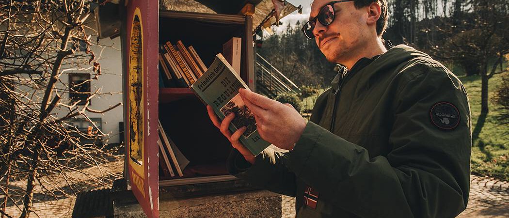 S-Bahn-Wandern: junger Mann mit Sonnenbrille liest Buch aus öffentlichem Bibliotheksschrank
